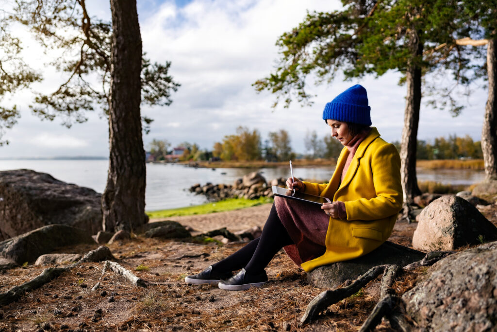 Keltatakkinen nainen istuu kivellä luonnossa ja pitelee tablettia ja sen kynää.