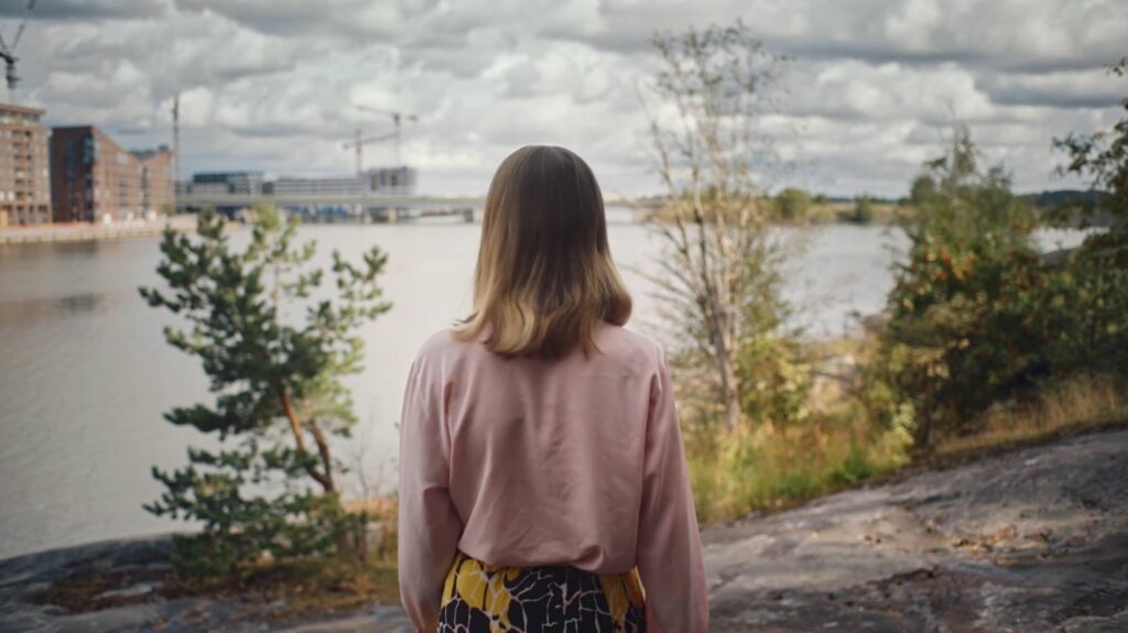 Nainen seisoo selkä kameraan päin ja katselee merelle. Sivuilla pienet puut.