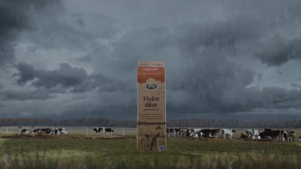 Arla Yhden tilan luomumaitopurkki seisoo nurmikolla synkän pilvisenä päivänä. Taustalla lehmiä laitumella.