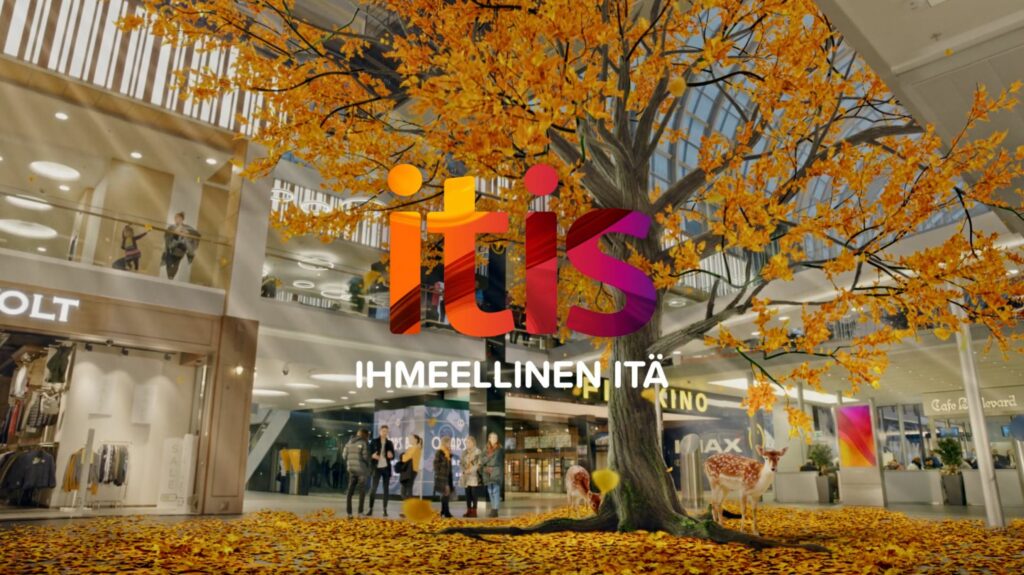 Itiksen logo. Taustalla kauppakeskus, jossa kasvaa syksyinen, lehtiä pudottava puu. itä
