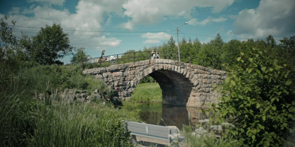 Kesäinen kuva kivisillasta joka ylittää pienen joen. Paljon virhreää metsää.