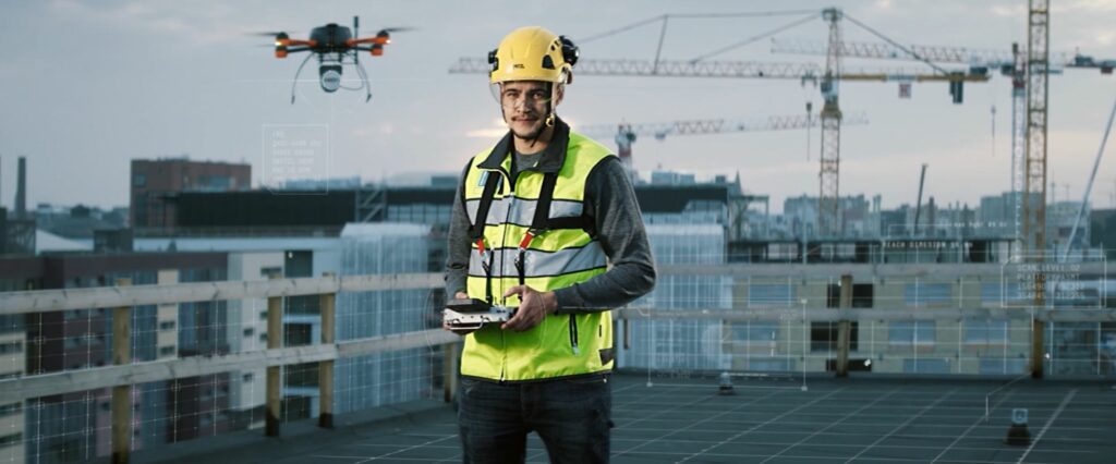 Mies keltaisissa suojavarusteissa pitelee drone ohjainta. Taustalla nostureita ja työmaata.