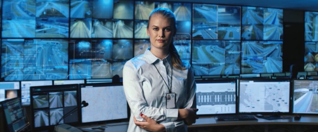 Kauluspaitainen nainen seisoo kädet ristissä. Taustalla useita näyttöjä, joissa tienäkymiä.