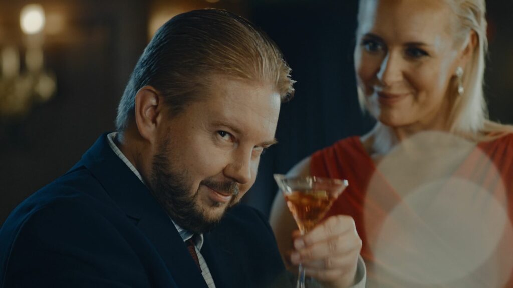 Lähikuva miehestä, joka katsoo oikealle ja nostaa drinkkilasia. Taustalla nainen juhlapukeissa.