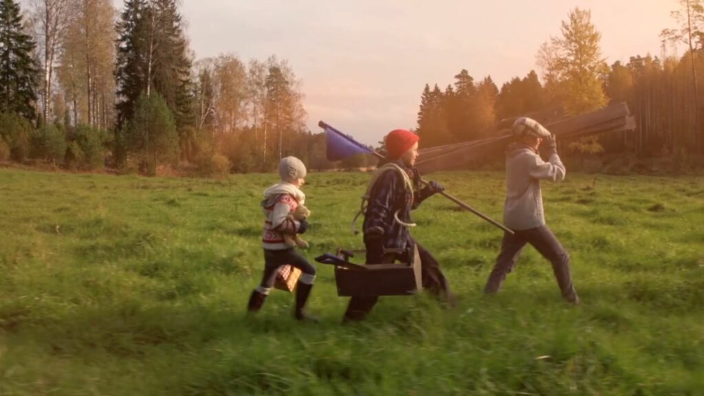 Kolme lasta kulkee pitkin vihreää niittyä kantaen rakennustarvikkeita. Taustalla metsää.