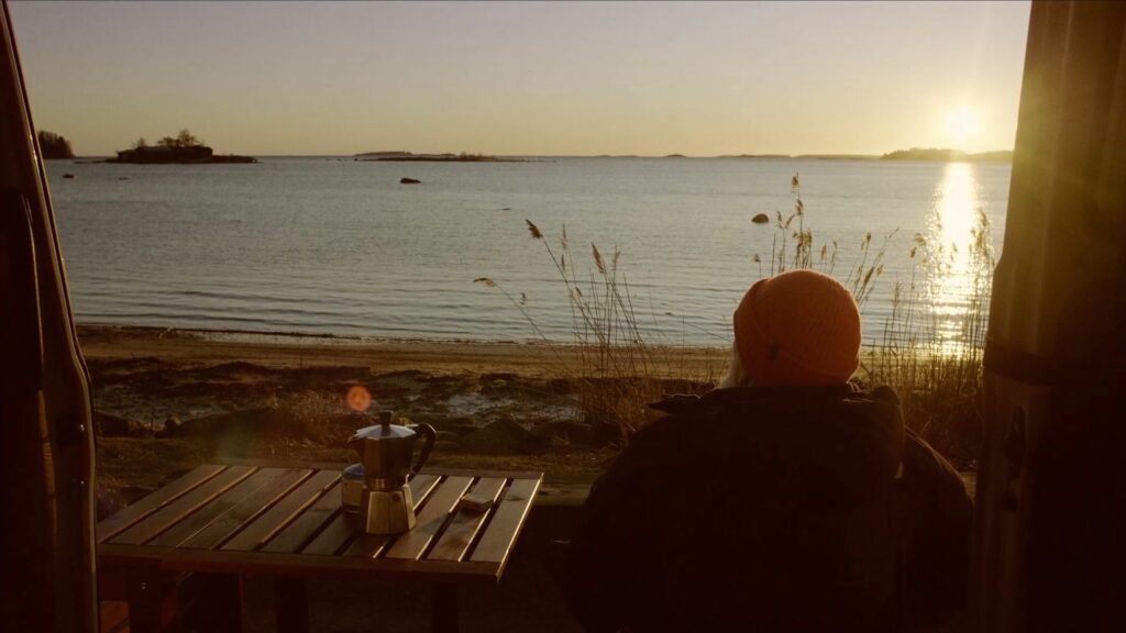 Ranta ja horisonttiin jatkuva vesi. Etualalla pöydän vieressä istuva mies katsomassa vettä päin.