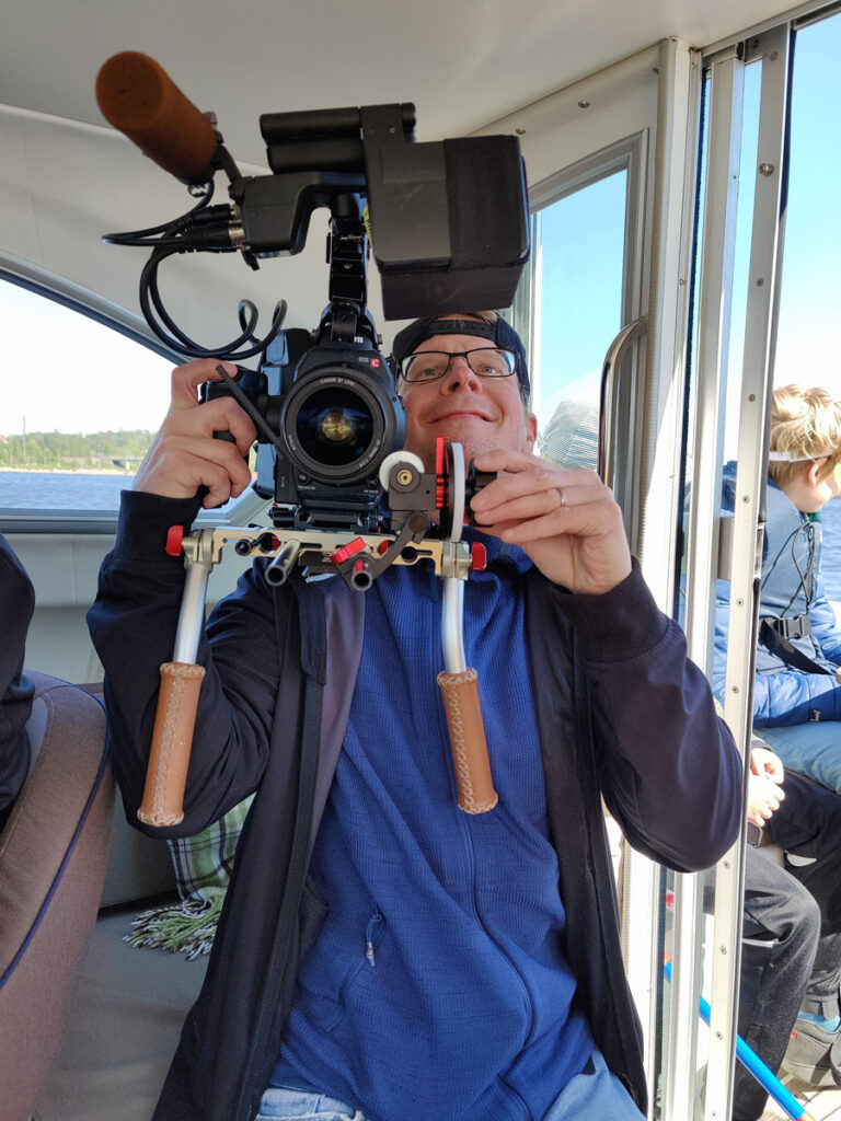Behind the Scenes. Videokuvaaja kuvaa hymyillen kohti kameraa.