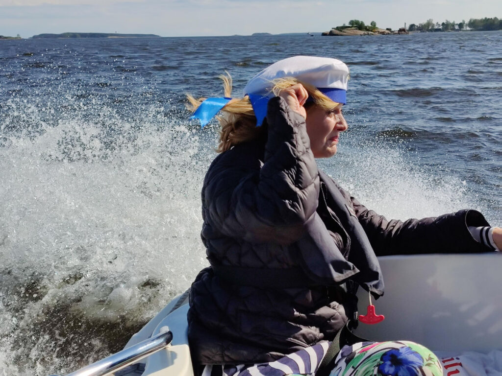 Sivukuva naisesta, joka istuu veneen kyydissä hattua pidellen. Vesi roiskuu ympärillä kovasta vauhdista.