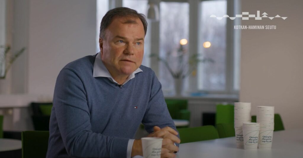 Mies istuu pöydän äärellä kahvikuppi edessään. Taustalla ikkuna ja huonekasvi.