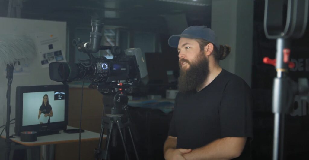 Behind the scenes, parrakas mies seisoo videokameran takana ja katsoo kameraan.
