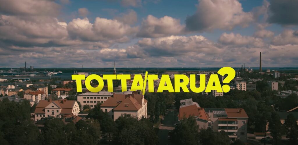 Paljon taloja, joissa punainen katto. Puita niiden seassa. Taivaalla paljon pilviä. Keltainen "totta/tarua" teksti keskellä.