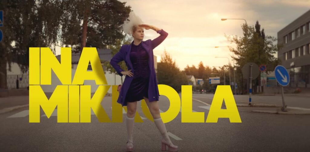 Ina Mikkola seisoo autotien keskellä ja tähyilee horisonttiin. Taustalla on puita ja aurinko laskee.