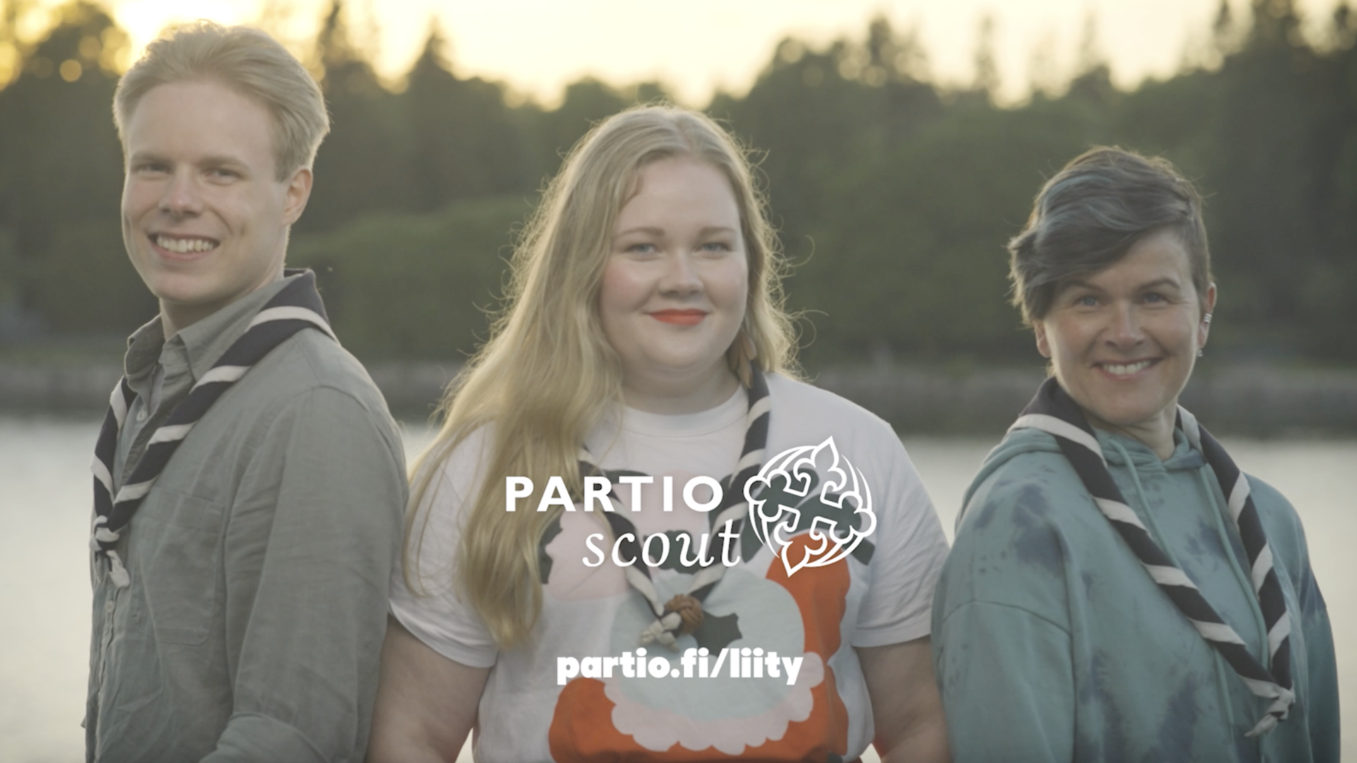 Kolme henkilöä katsovat suoraan kameraan kauniissa kesäillassa meri takana.